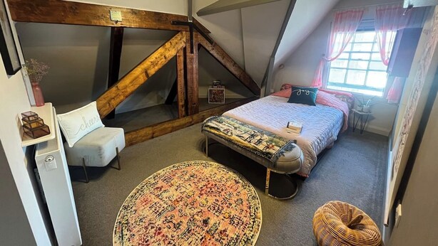 A loft bedroom with a sloped ceiling, a bed with a patterned bedspread, a round rug, a small round table, and a chair. - Gracious Apartment in Historic Sheffield Georgian Quarter (Sheffield City Centre)