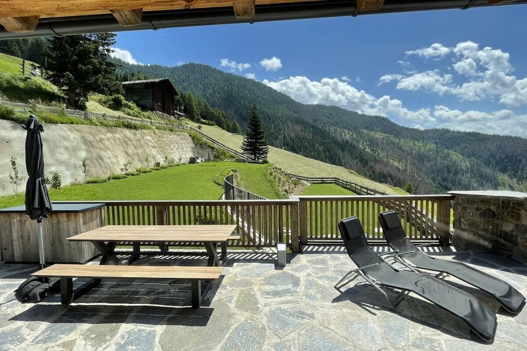 A patio with a wooden table and two lounge chairs, overlooking a mountainous landscape with a wooden cabin.