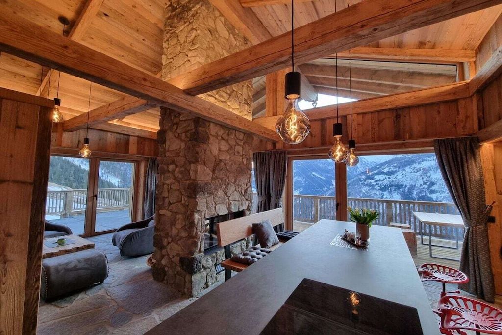 Ein Holzinnenraum mit Kamin aus Stein, einer Sitzecke und großen Fenstern mit Blick auf einen verschneiten Berg.