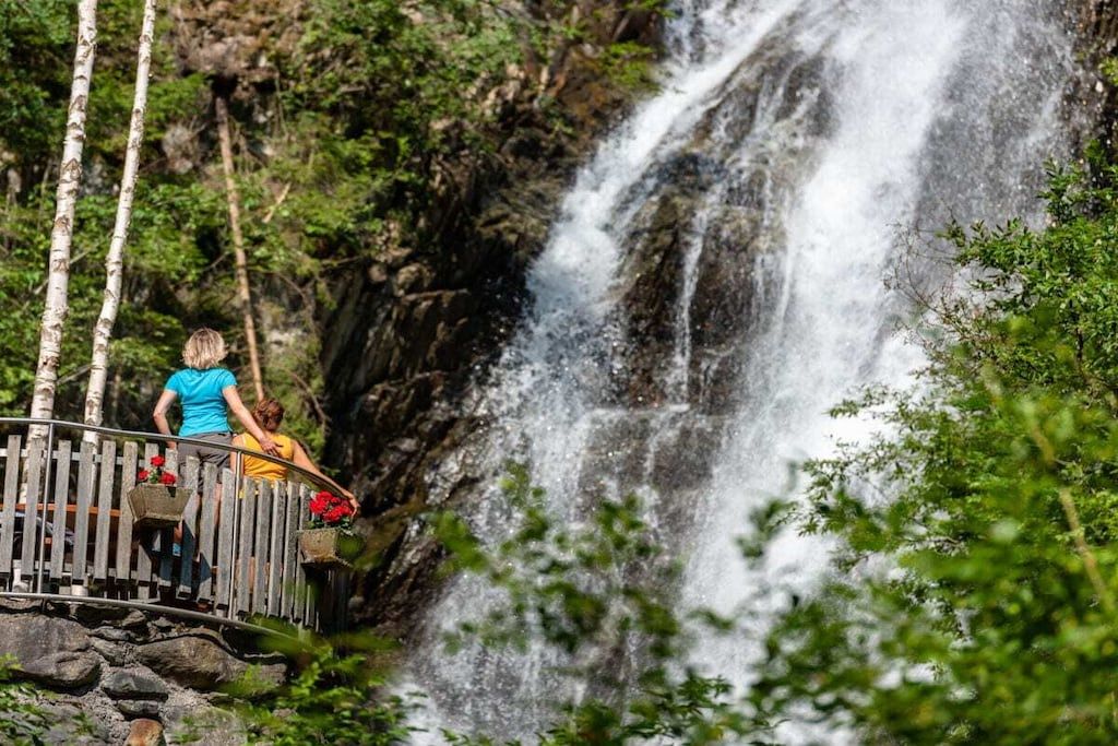 Un pont en bois, doté de garde-corps, mène à une cascade entourée de verdure.