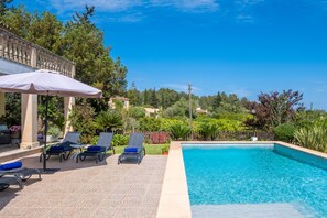 Ein Swimmingpool mit Liegestühlen und einem Sonnenschirm, umgeben von viel Grün und blauem Himmel.