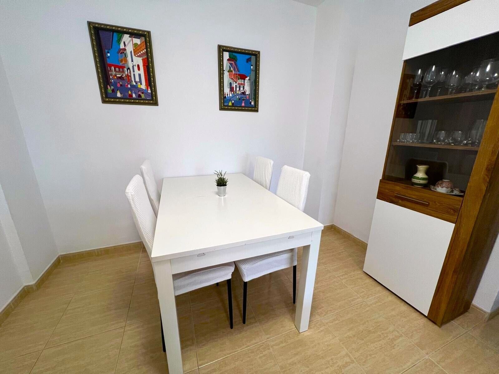 Een witte eettafel met vier stoelen, een kleine potplant en twee ingelijste kunstwerken aan de muur.