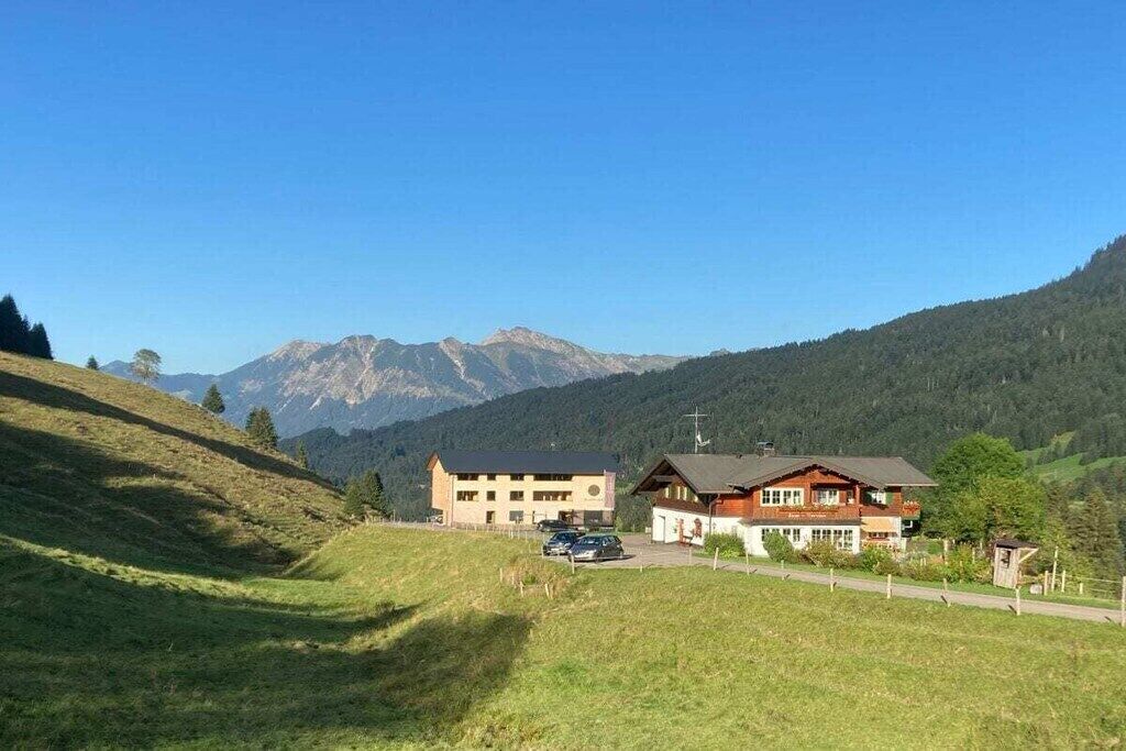 Ein zweistöckiges Gebäude mit Parkplatz davor, umgeben von einer Wiese und Bergen im Hintergrund.