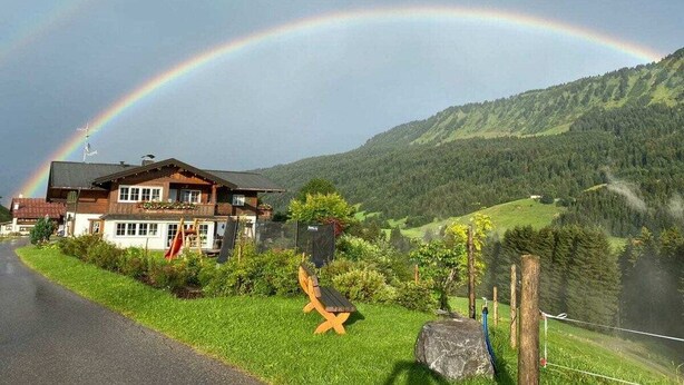 A building with a large wooden structure, a flag, and a bench in a rural setting with a mountain backdrop. - Apartment in Riezlern with swimming pool (Riezlern)