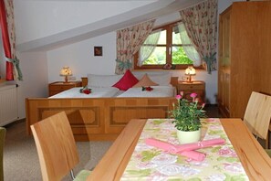 A bedroom with a wooden bed, two bedside tables with lamps, a window with floral curtains, and a table with a potted plant.
