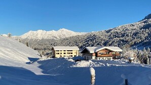 A snow-covered mountain landscape with two buildings, one with a light facade and the other with a dark wooden facade, both with snow-covered roofs. - Apartment in Riezlern with swimming pool (Riezlern)
