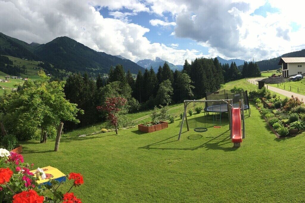 Ein grünes Freigelände mit Schaukel und Rutsche, umgeben von Bäumen und Bergen im Hintergrund.