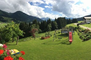 A grassy outdoor area with a swing set and a slide, surrounded by trees and mountains in the background.