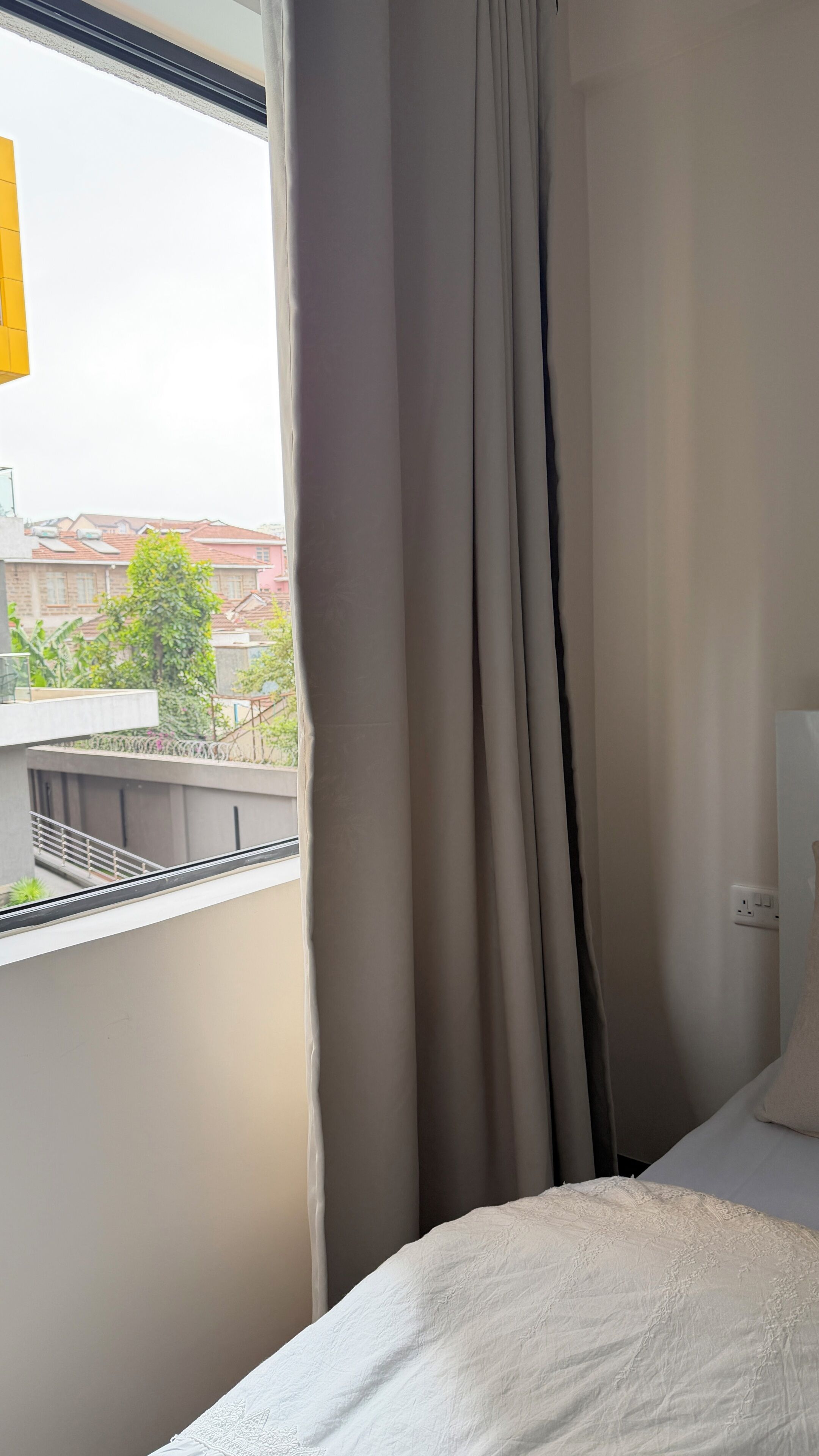 A hotel room with a bed, a window with curtains, and a view of buildings and greenery outside.