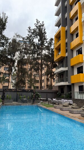 Una piscina con zona de solárium empedrado, rodeada por un edificio de varios pisos con balcones.