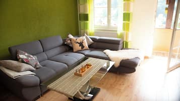Ein modernes Wohnzimmer mit einer grauen Eckcouch, einem Glastisch und Blick ins Freie durch ein großes Fenster.