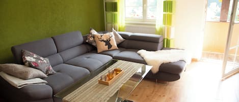 Un soggiorno moderno con un divano angolare grigio, un tavolino in vetro e vista sull'esterno attraverso un'ampia finestra.