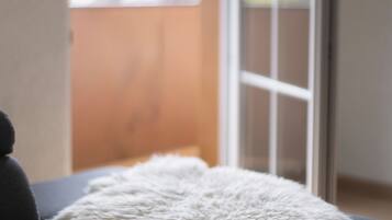 Ein Zimmer mit einem Bett, einer weichen, weißen Decke und Blick auf einen Balkon mit Blumen.