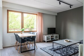 Ein modernes Wohnzimmer mit Essbereich, Fernseher und einem großen Fenster mit Blick auf Bäume.