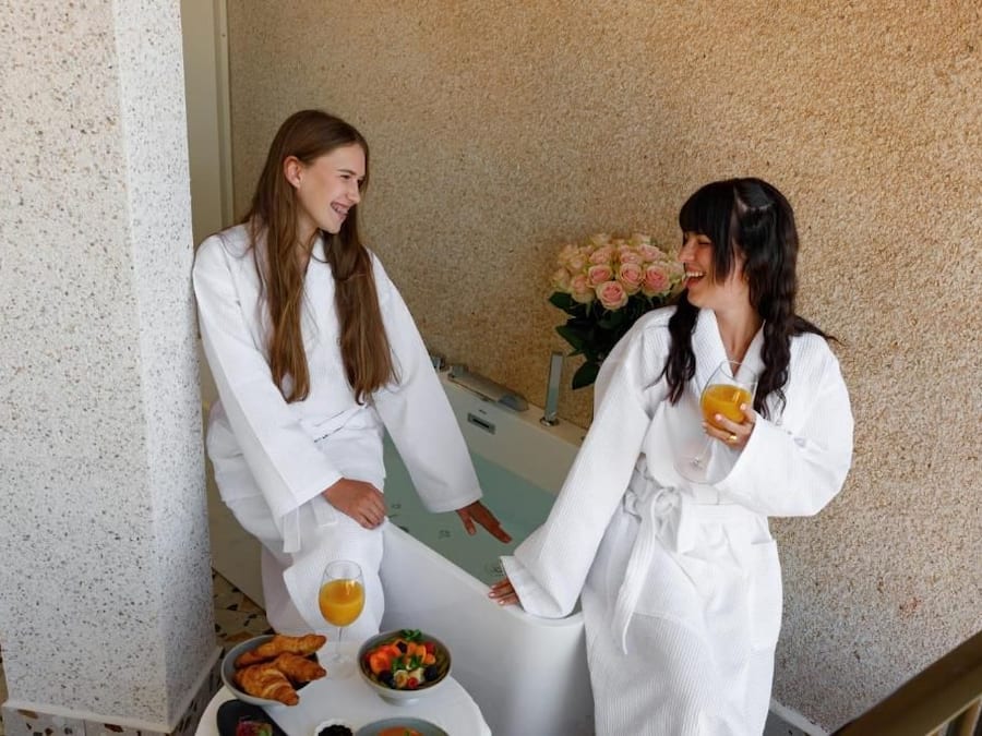 Dos personas vestidas de blanco de pie junto a una mesa con alimentos y bebidas para el desayuno.