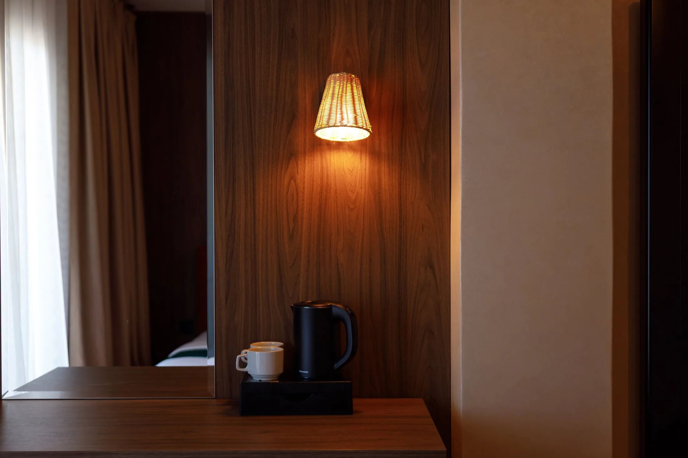 A wooden bedside table with a black electric kettle and a white mug, a wall-mounted lamp, and a wooden panel wall.
