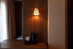 Una mesita de noche de madera con una tetera eléctrica negra y una taza blanca, una lámpara de pared y un panel de madera en la pared.