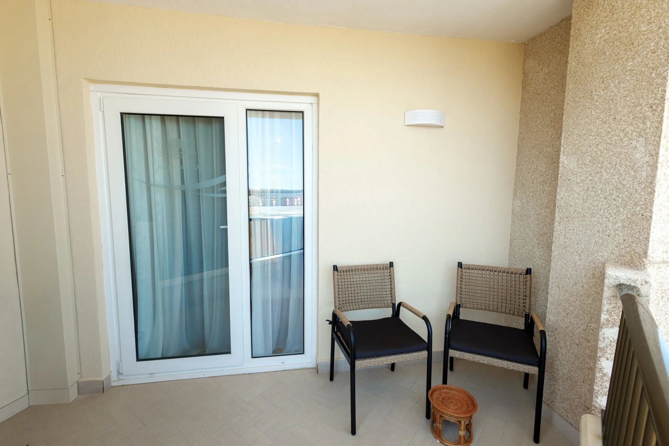 Un balcon avec deux fauteuils en osier, un petit tabouret en osier et une porte-fenêtre à glissière avec des rideaux.