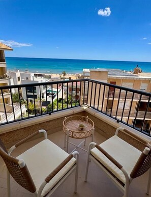 A balcony with two wicker chairs and a small round table, overlooking a beach and cityscape.