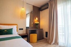 A hotel room with a bed, a bedside table, a small stool, a mirror, a wall-mounted light, and a window with curtains.