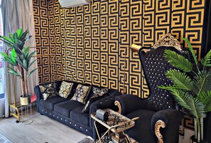Ein modernes Wohnzimmer mit einer Wand in Musteroptik, einem dunklen Sofa mit Zierkissen, einem kleinen Beistelltisch und einer großen Kübelpflanze.