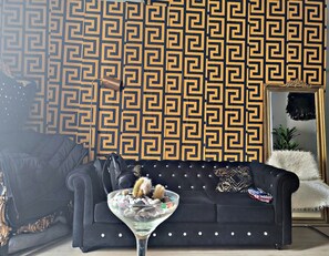Ein Zimmer mit Wandmuster, einer schwarzen Couch mit dekorativen Kissen, einem Glastisch mit Steinen und einem Spiegel mit goldener Rahmung.