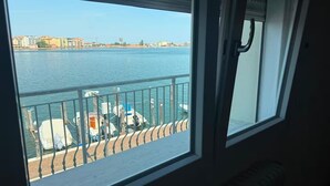 Vista desde una ventana sobre aguas con barcos amarrados, barandilla de balcón y edificios a lo lejos.