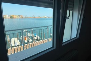 Blick aus einem Fenster auf ein Gewässer mit dort vor Anker liegenden Booten, einem Balkongeländer und Gebäuden in der Ferne.
