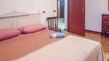 Ein Bett mit einer rosa Bettdecke und Kissen, ein blaues Handtuch und ein Holzstuhl in einem Zimmer mit weißer Wand und Holztür.