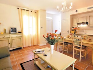 A living room with a television, a dining table with chairs, a vase with flowers, and a kitchen area with cabinets and a chandelier. - Cabin House for 4 in Provence (Fayence)