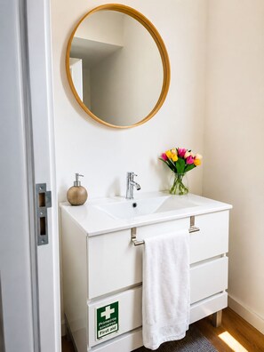 Ein Badezimmer mit einem weißen Waschtisch, einem runden Spiegel, einem Waschbecken, einem Handtuch, einem Erste-Hilfe-Schild und einer Vase mit Blumen.