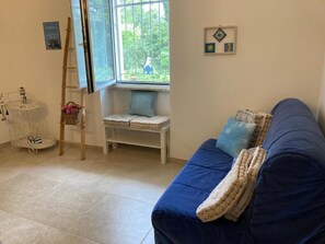Una habitación con un sofá azul, una mesita blanca, un ventanal con vistas a la vegetación y una escalera apoyada en la pared.