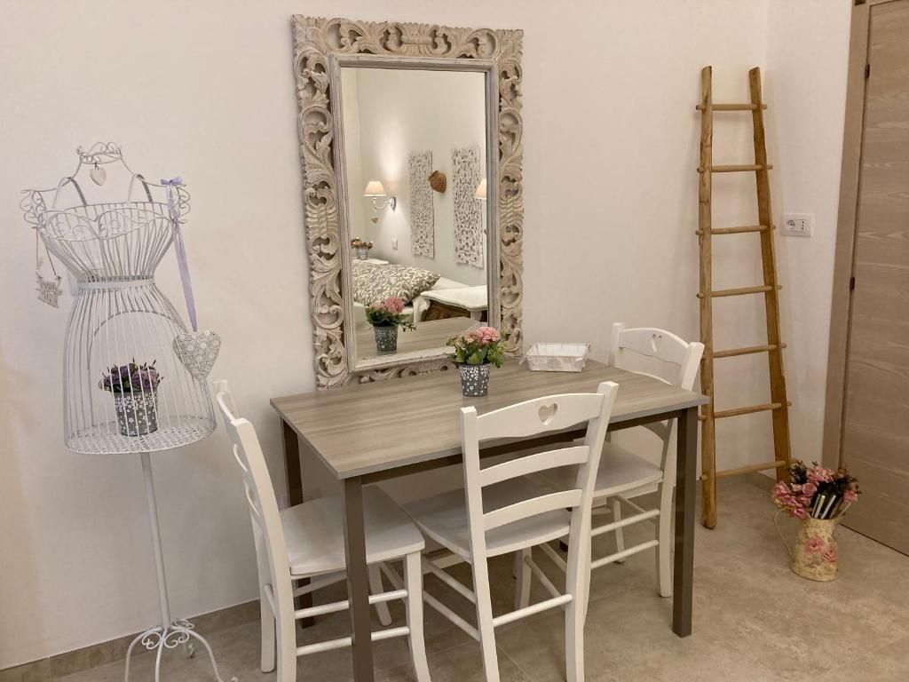 Una habitación con una mesa de comedor de madera, sillas blancas, un espejo con un marco ornamentado, un soporte de metal blanco con un objeto decorativo y una escalera de madera.