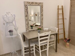 Una habitación con una mesa de comedor de madera, sillas blancas, un espejo con un marco ornamentado, un soporte de metal blanco con un objeto decorativo y una escalera de madera.