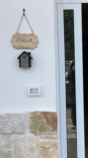 Un letrero de madera con el nombre 'PERLA' y un comedero para pájaros en una pared, una puerta con un panel de vidrio y una pared de piedra.
