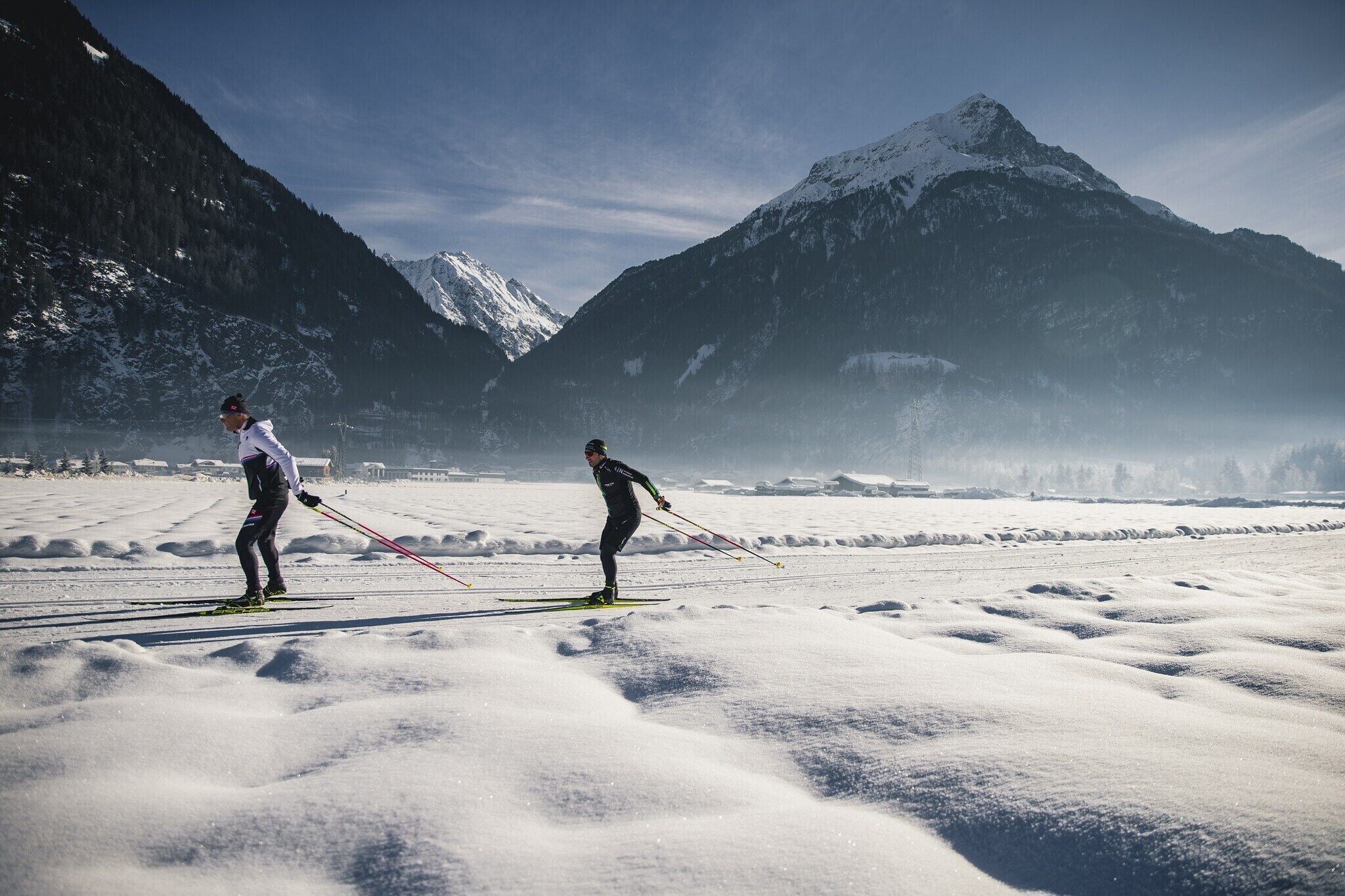 Twee skiërs op een besneeuwde berg, met een heldere lucht en een berg op de achtergrond.