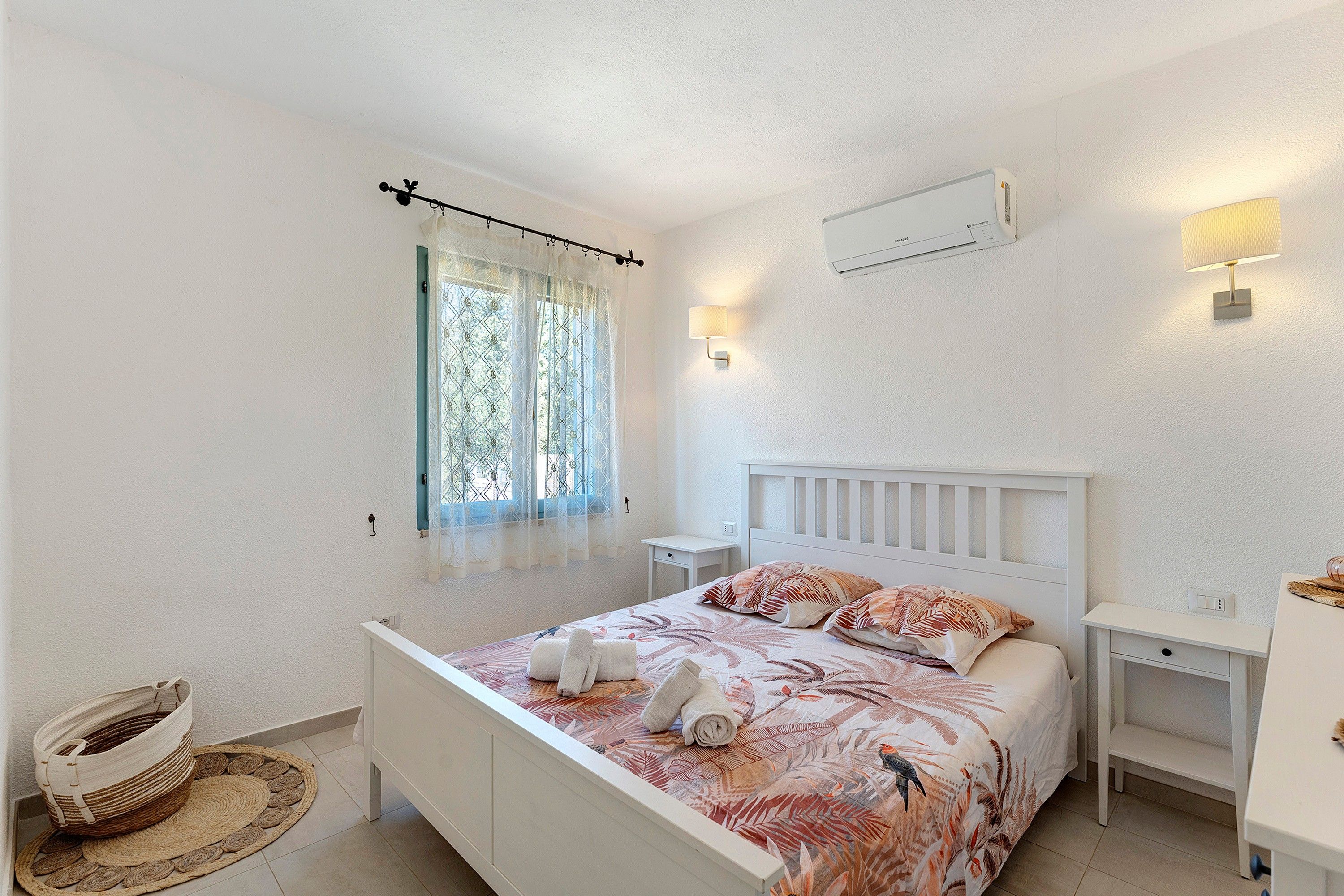 Un dormitorio con cama, mesitas de noche, una ventana con cortinas, un aire acondicionado instalado en la pared y una cesta tejida.