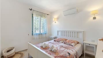 Un dormitorio con cama, mesitas de noche, una ventana con cortinas, un aire acondicionado instalado en la pared y una cesta tejida.