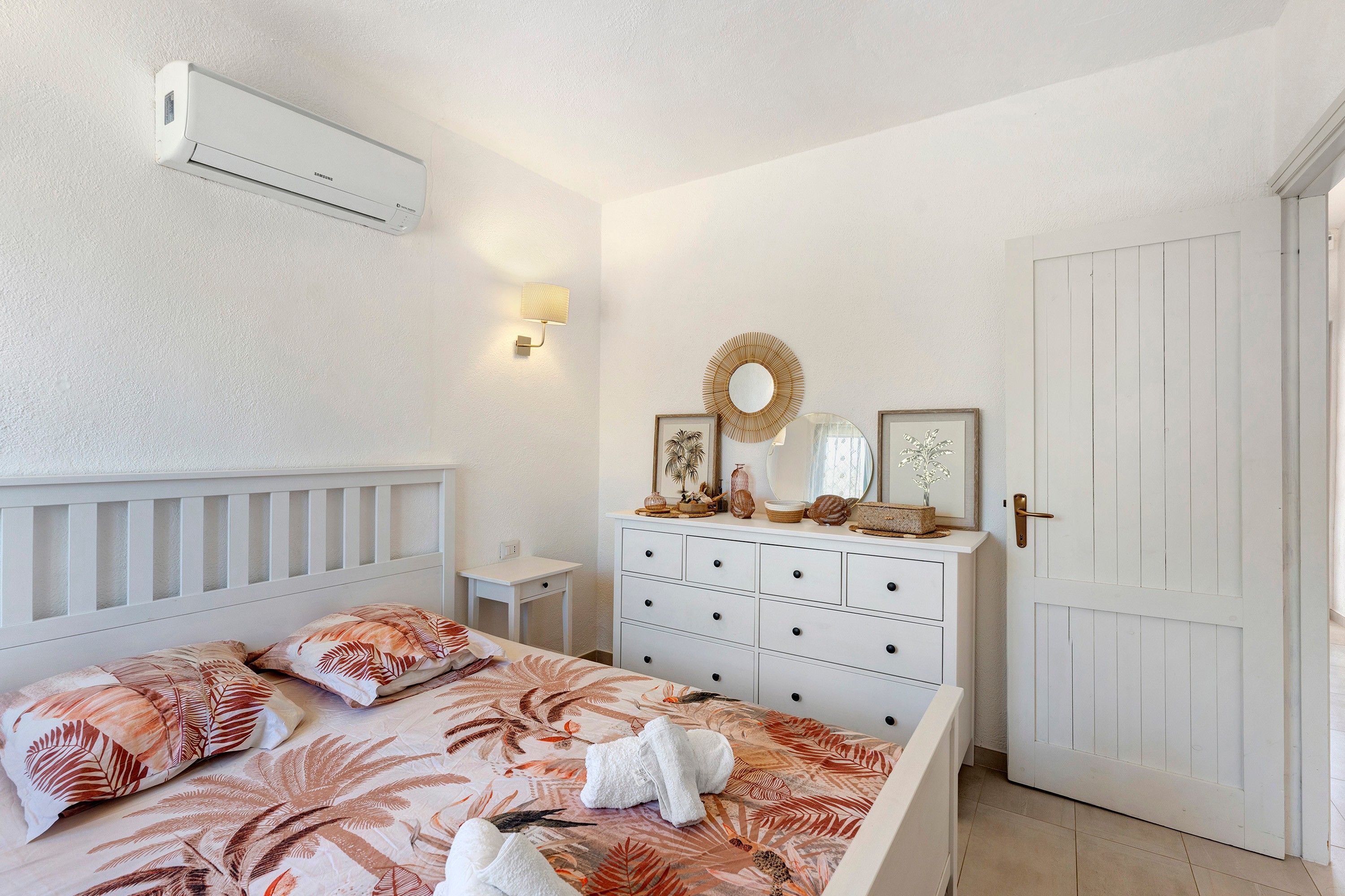 Un dormitorio con cama, cómoda, aire acondicionado de pared, una mesita y una puerta.