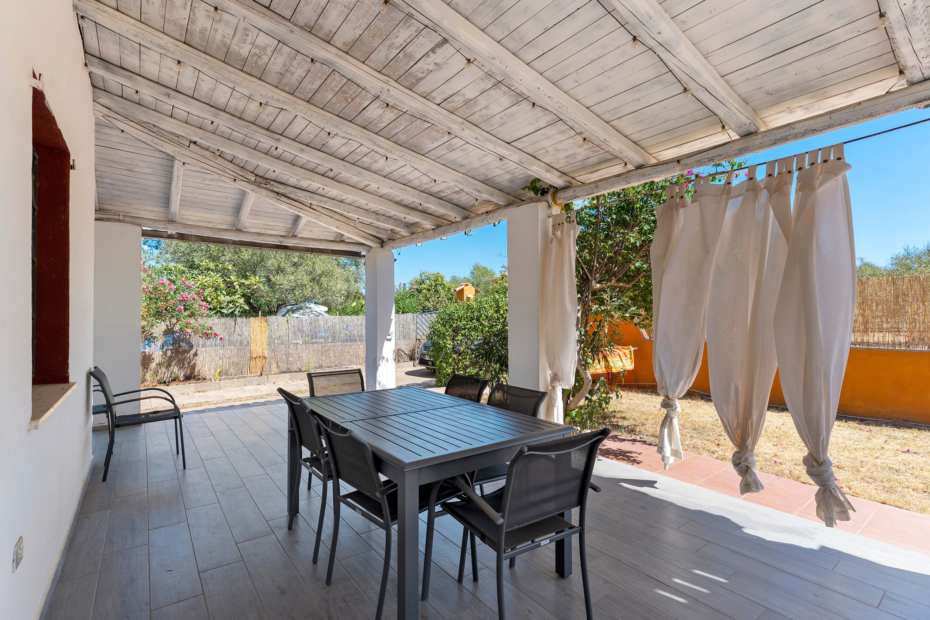 Un patio techado al aire libre con una mesa y sillas, techo de madera y vista del jardín circundante.