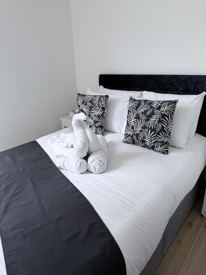 A neatly made bed with white bedding and black leaf-patterned pillows, a dark headboard, and a white nightstand with a lamp. - Modern 2BR/2BA | Private Balcony & Free Parking (Hertfordshire)
