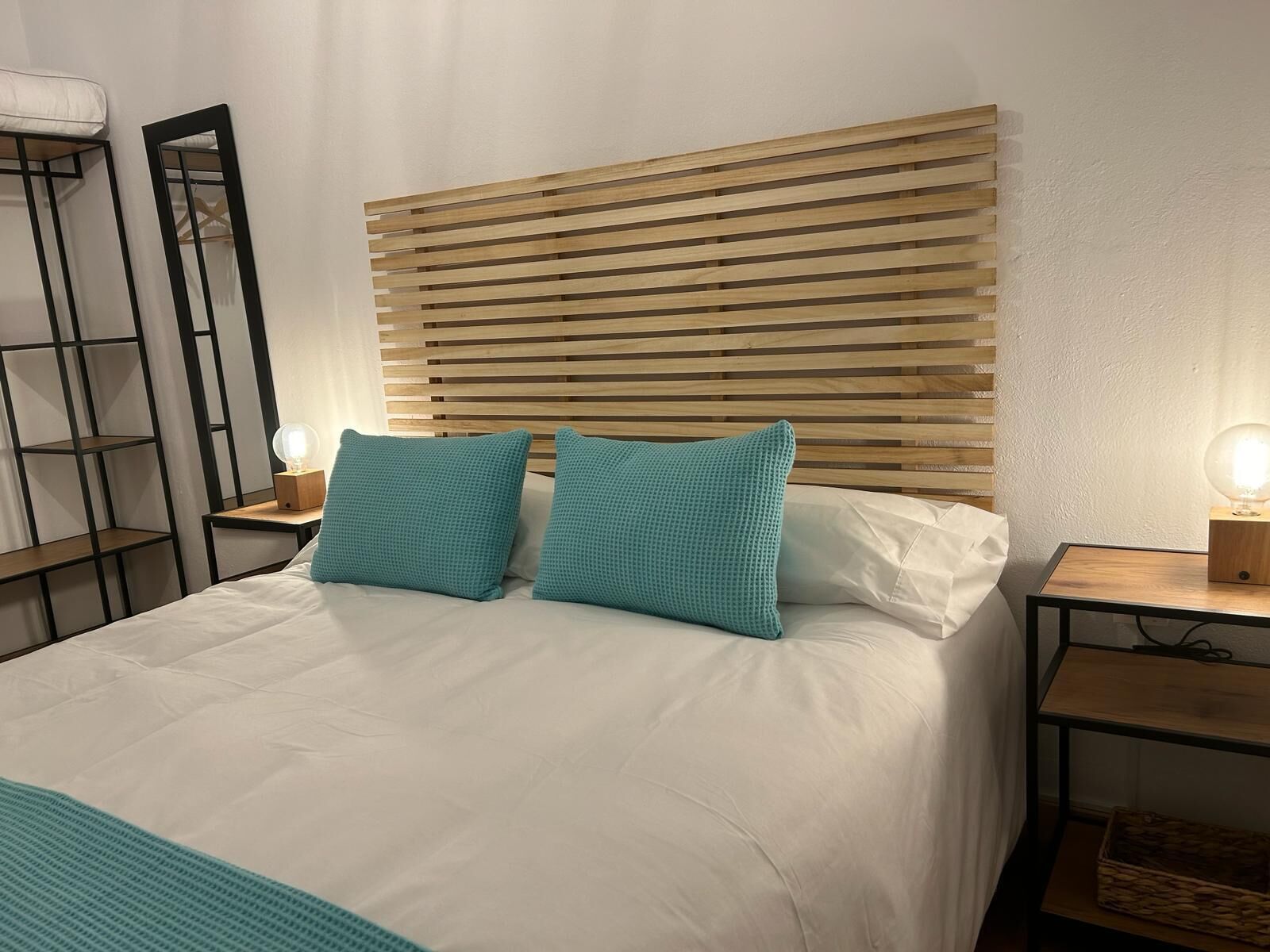 A neatly made bed with two pillows, a wooden slatted headboard, a bedside table with a lamp, and a small shelf with a basket.