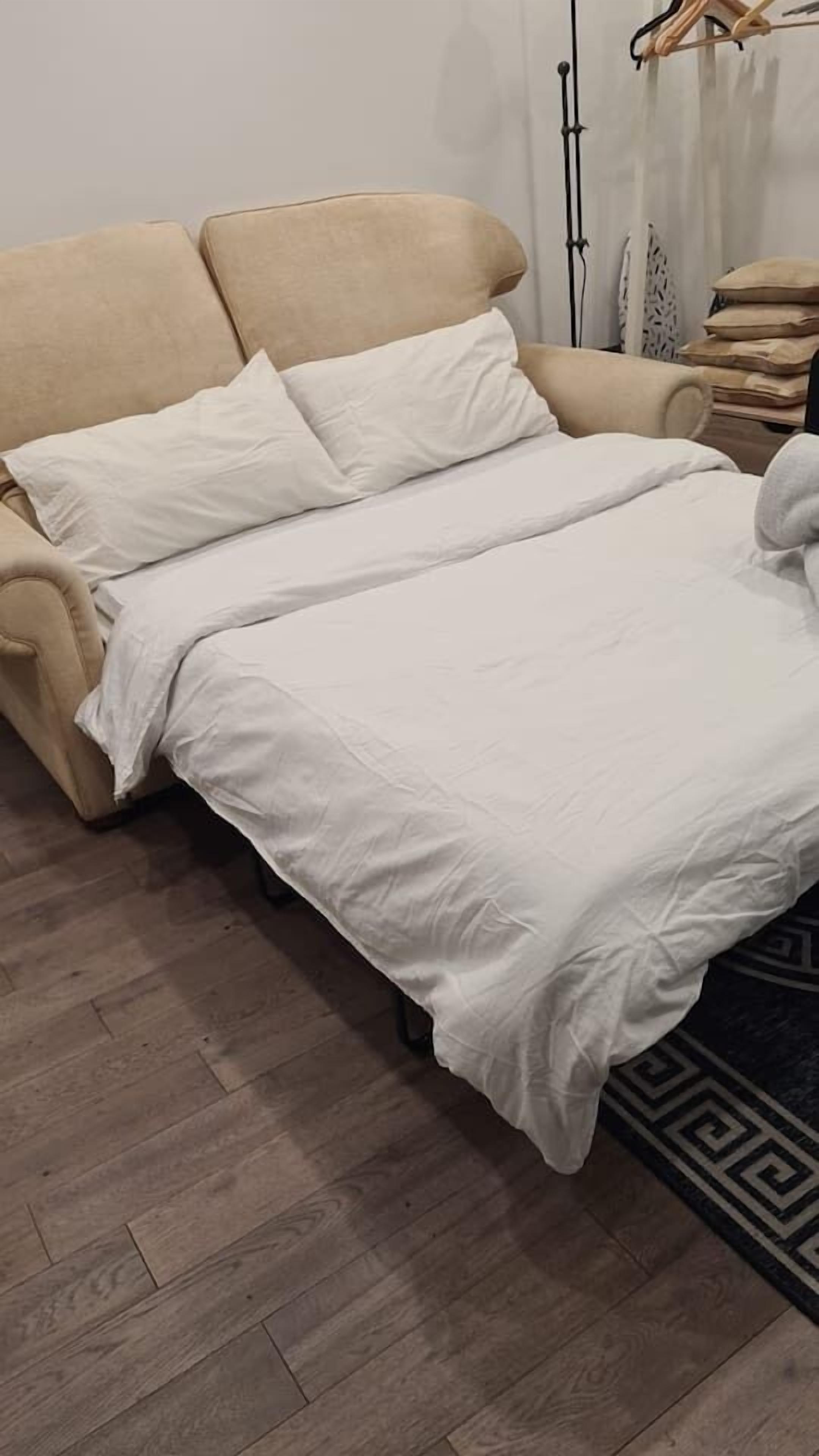A beige sofa with a white bedspread and pillows in a room with wooden flooring and a patterned rug.