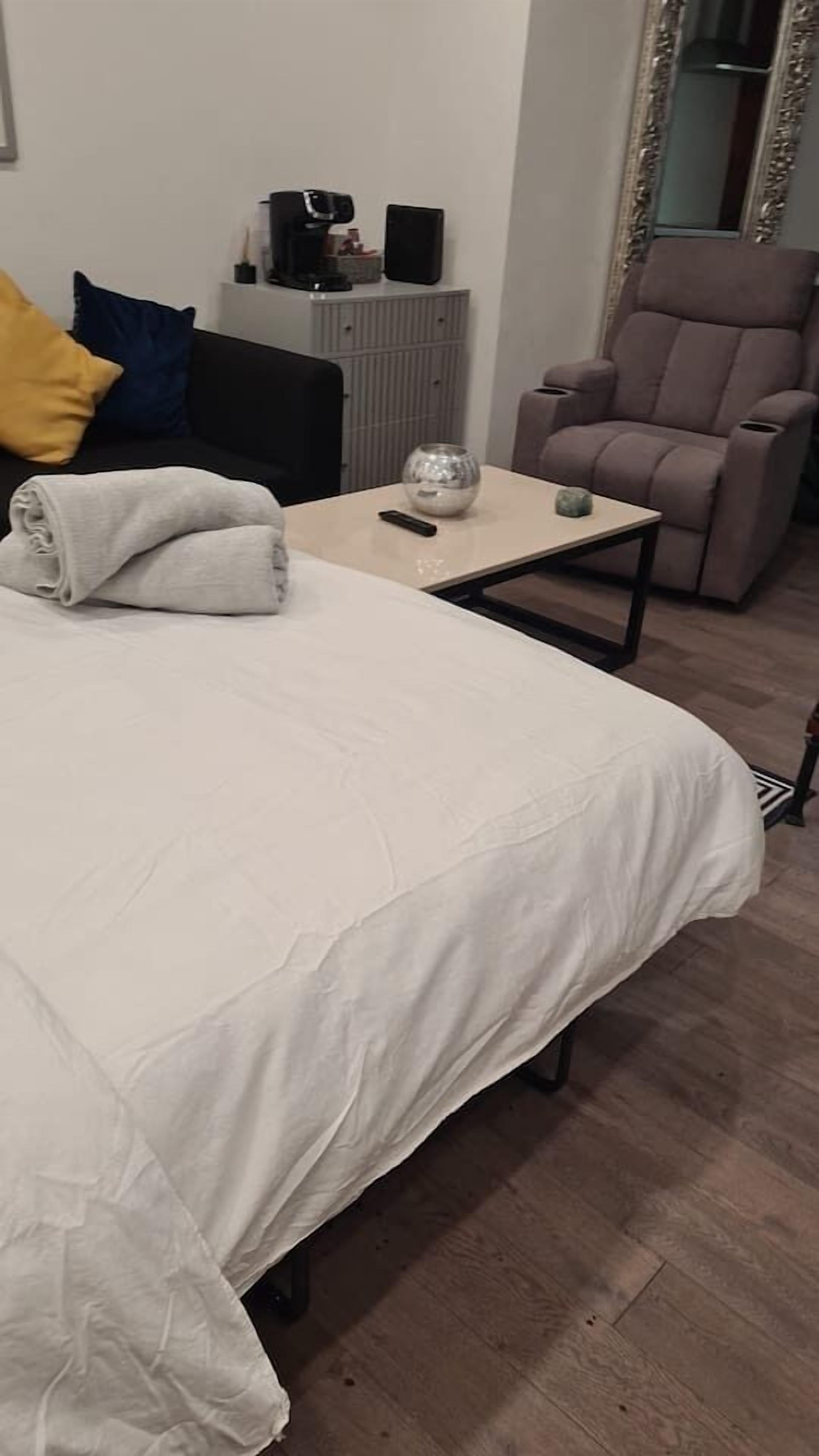 En pænt redt seng med et hvidt dynbetræk, et lille bord med en fjernbetjening, en kaffemaskine og en stol i et rum med trægulv og en radiator.