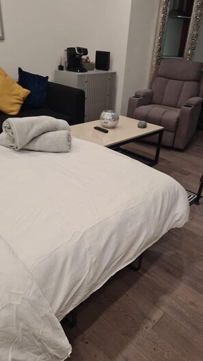 A neatly made bed with a white bedspread, a small table with a remote control, a coffee maker, and a chair in a room with wooden flooring and a radiator.