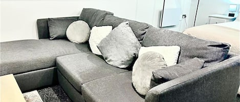 A modern living room with a grey sectional sofa, multiple cushions, a black floor lamp, and a white wall.