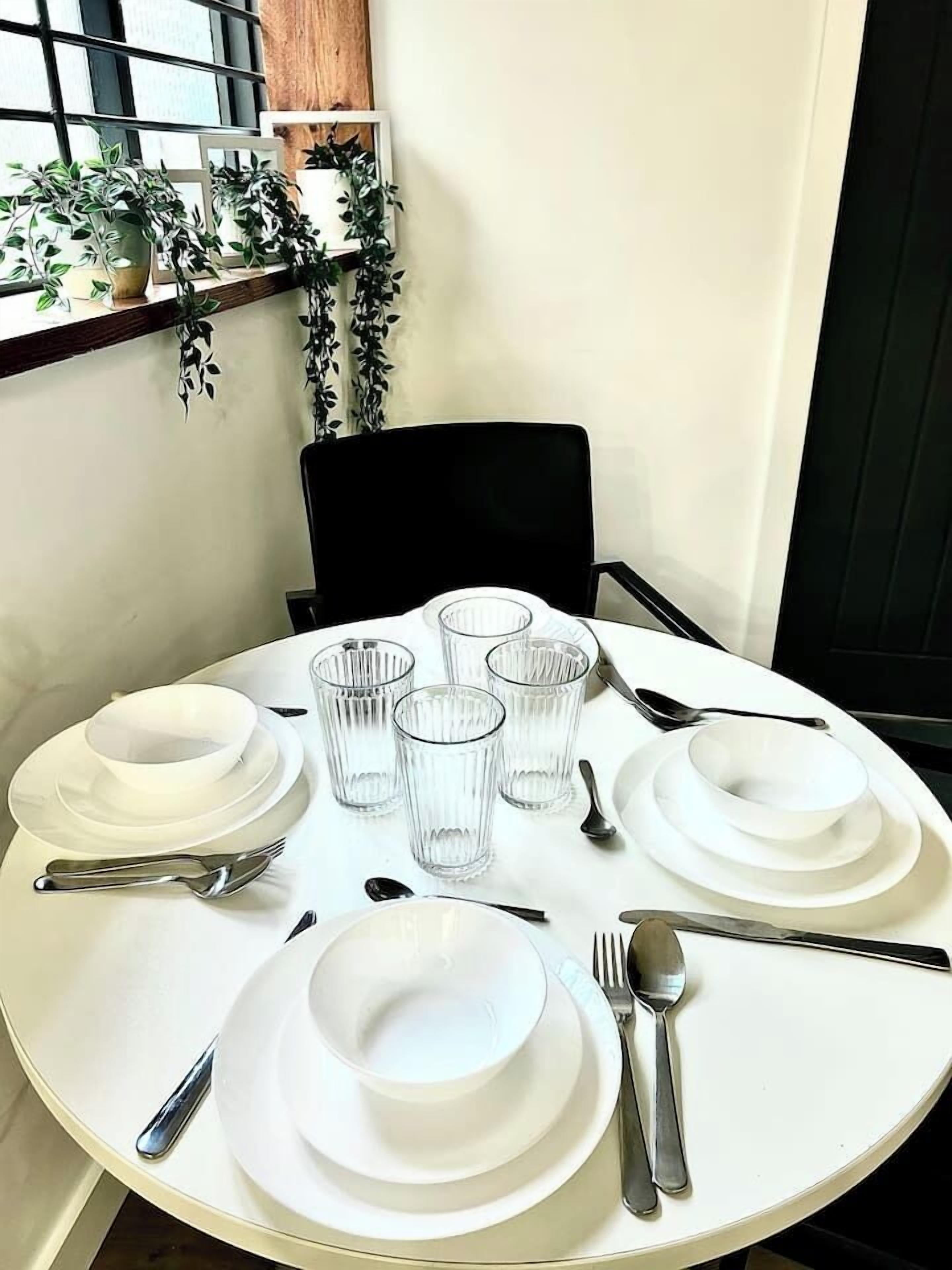 A round dining table set with white plates, glasses, and cutlery, positioned near a window with plants.