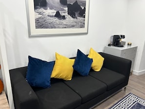En svart soffa med blå och gula kuddar, ett inramat svartvitt fotografi på väggen och en kaffemaskin på en vit bänkskiva.