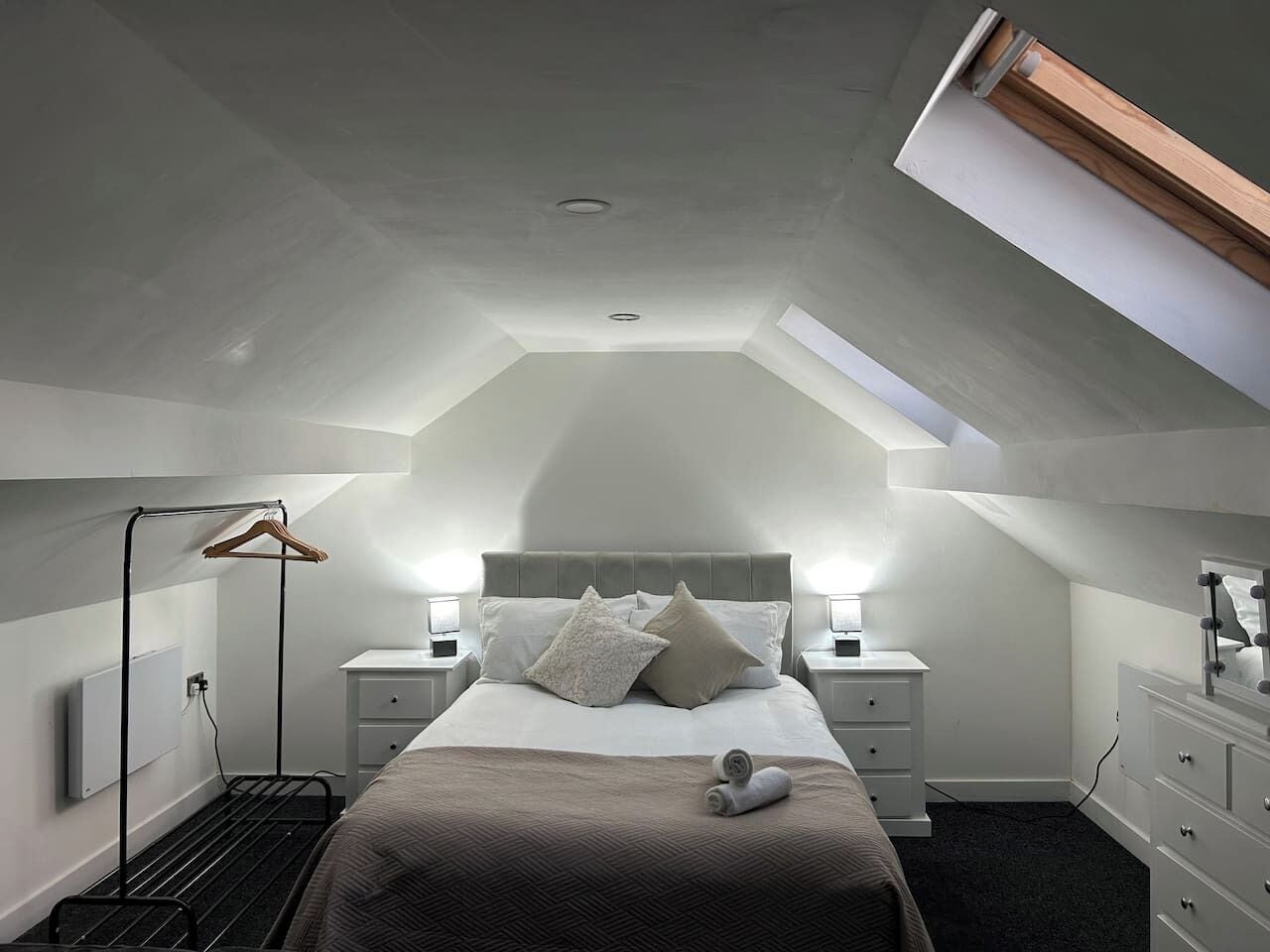 A bedroom with a sloped ceiling, a bed with pillows, bedside tables, a hanging clothes rack, and a dresser with a mirror.