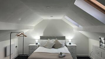 A bedroom with a sloped ceiling, a bed with pillows, bedside tables, a hanging clothes rack, and a dresser with a mirror.